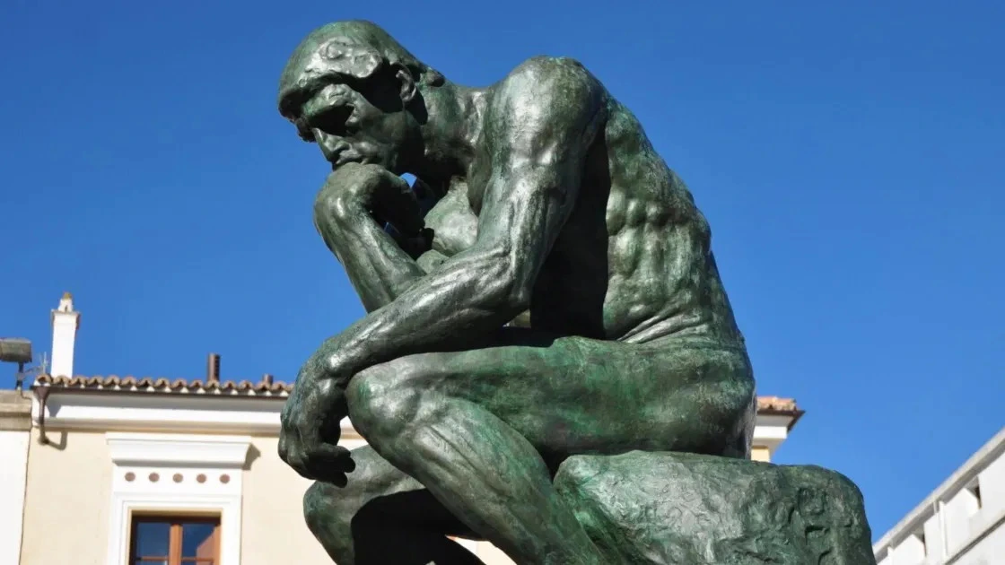 Fotografia da escultura de bronze 'O Pensador' de Rodin, mostrando um homem sentado em profunda contemplação contra um céu azul.